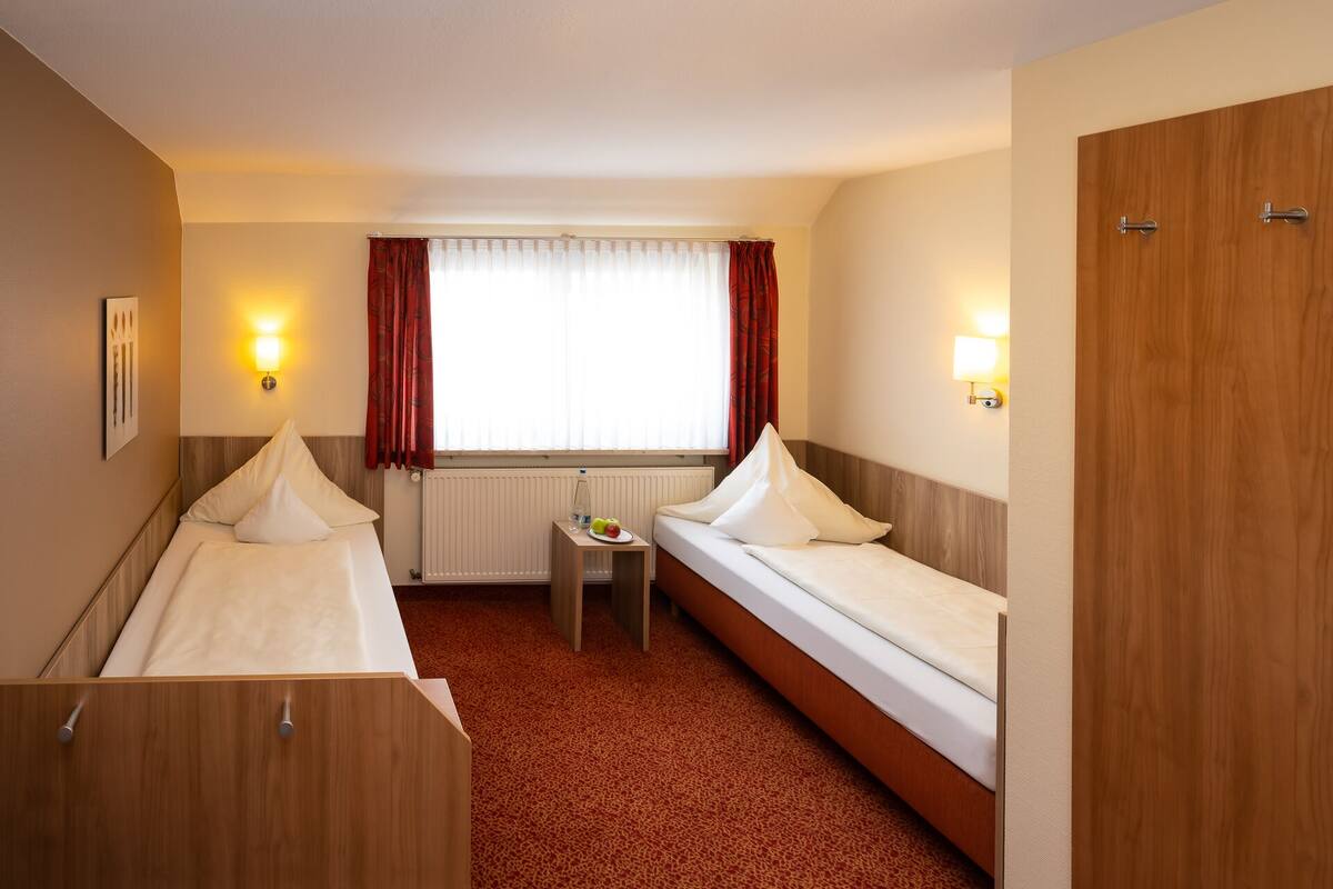 Une chambre d’hôtel avec deux lits simples, une petite table sur laquelle se trouve une assiette de fruits, un radiateur et deux lampes fixées au mur.