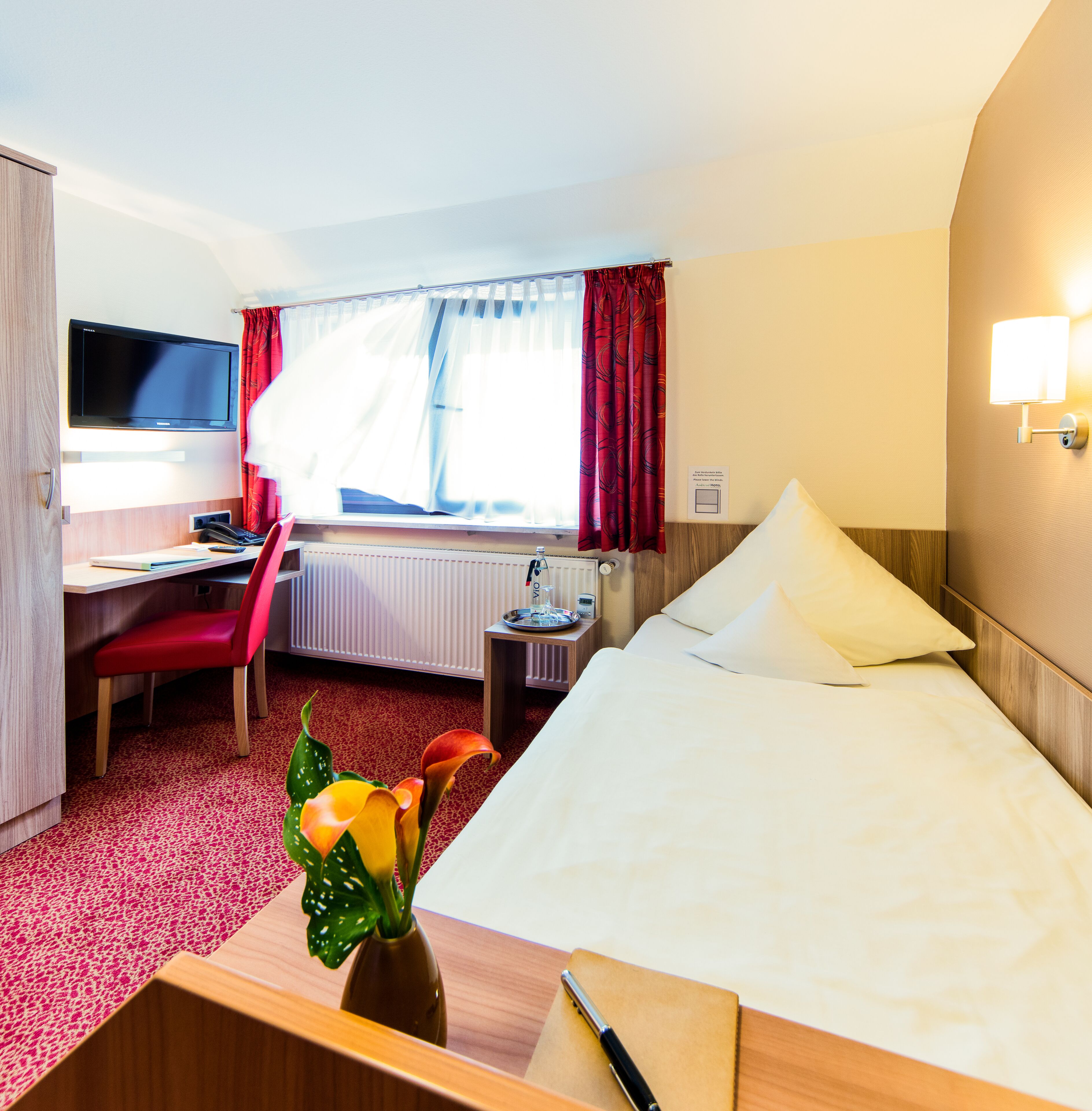 Habitación de hotel con una cama, un escritorio y una silla, un televisor, una ventana con cortinas y un jarrón con flores.