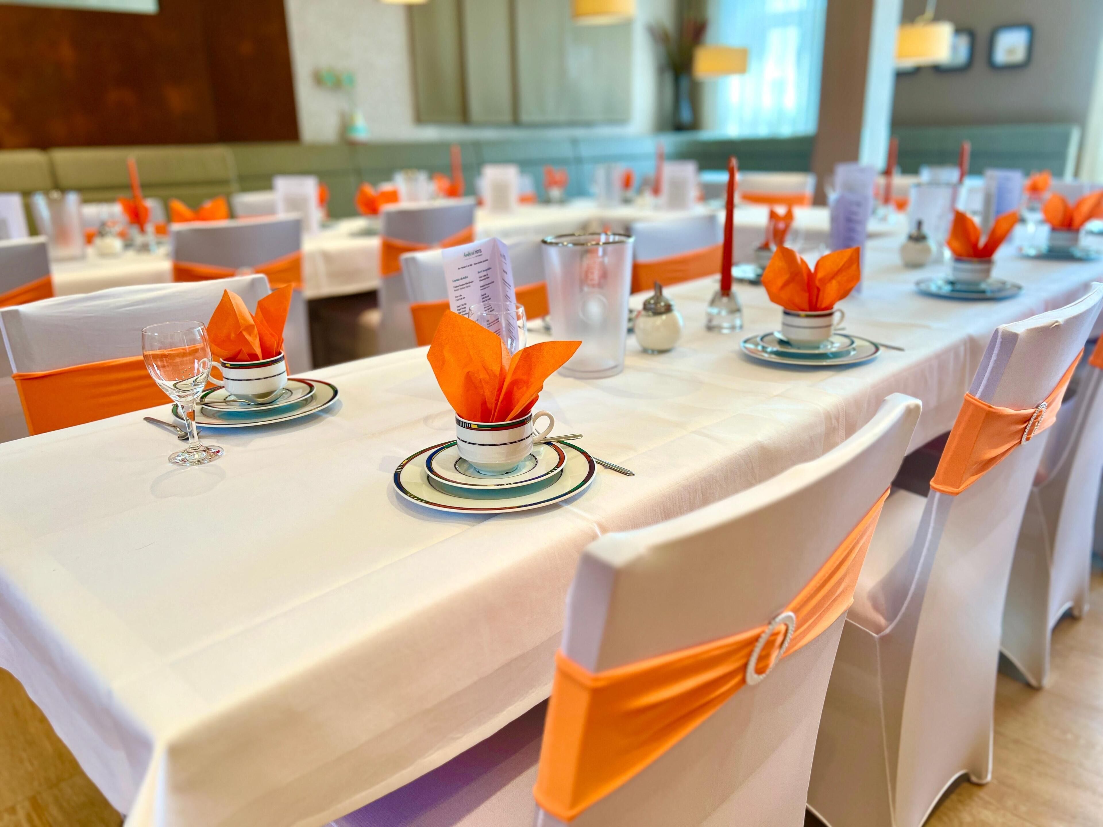 Un comedor con mesas dispuestas para el servicio, sillas con cintas naranjas y un atril para el menú.