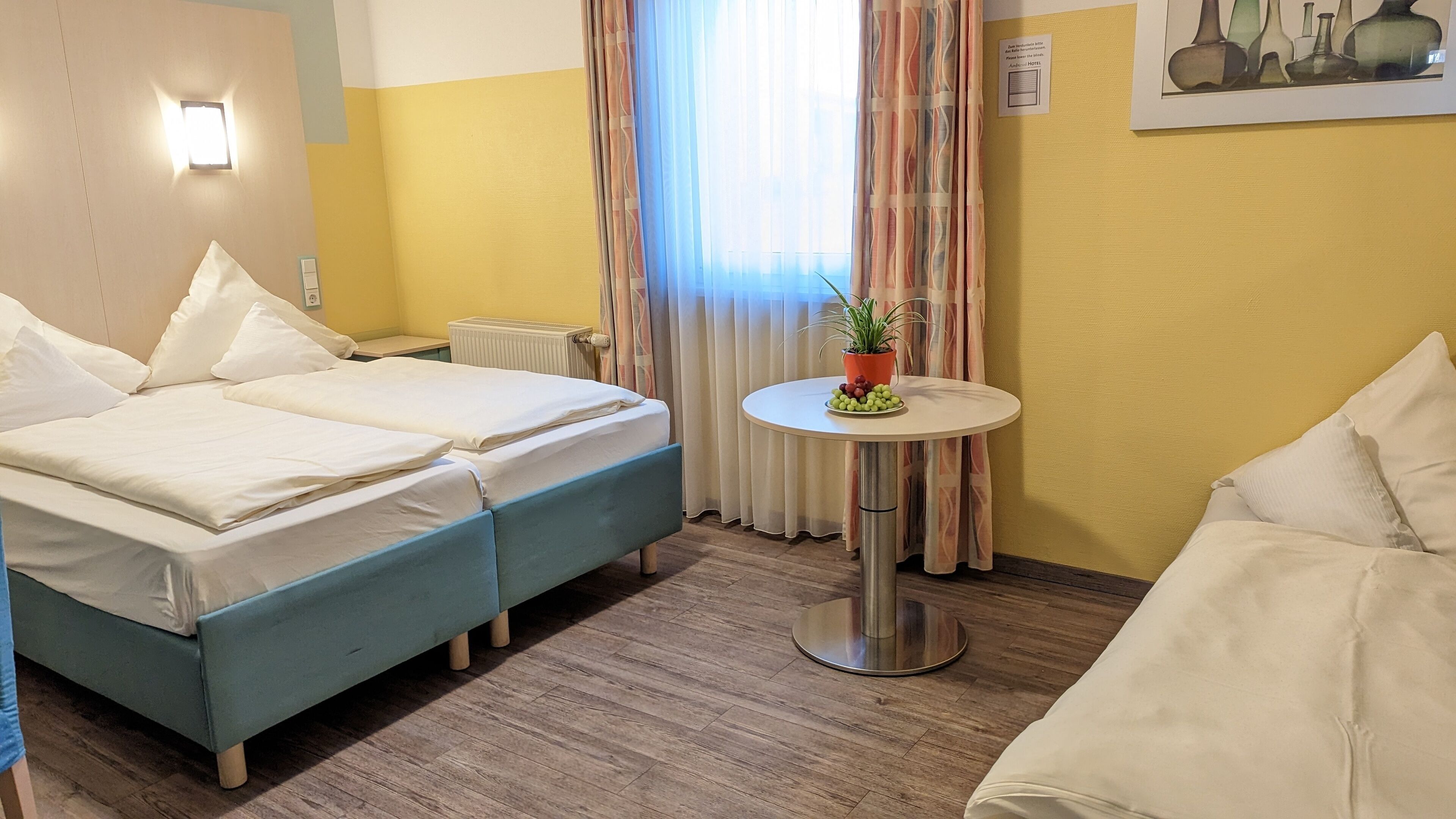 Hotellihuone, jossa on parivuode, pieni pyöreä pöytä, jonka päällä on kasvi ja hedelmiä, patteri ja ikkuna, jossa on verhot.