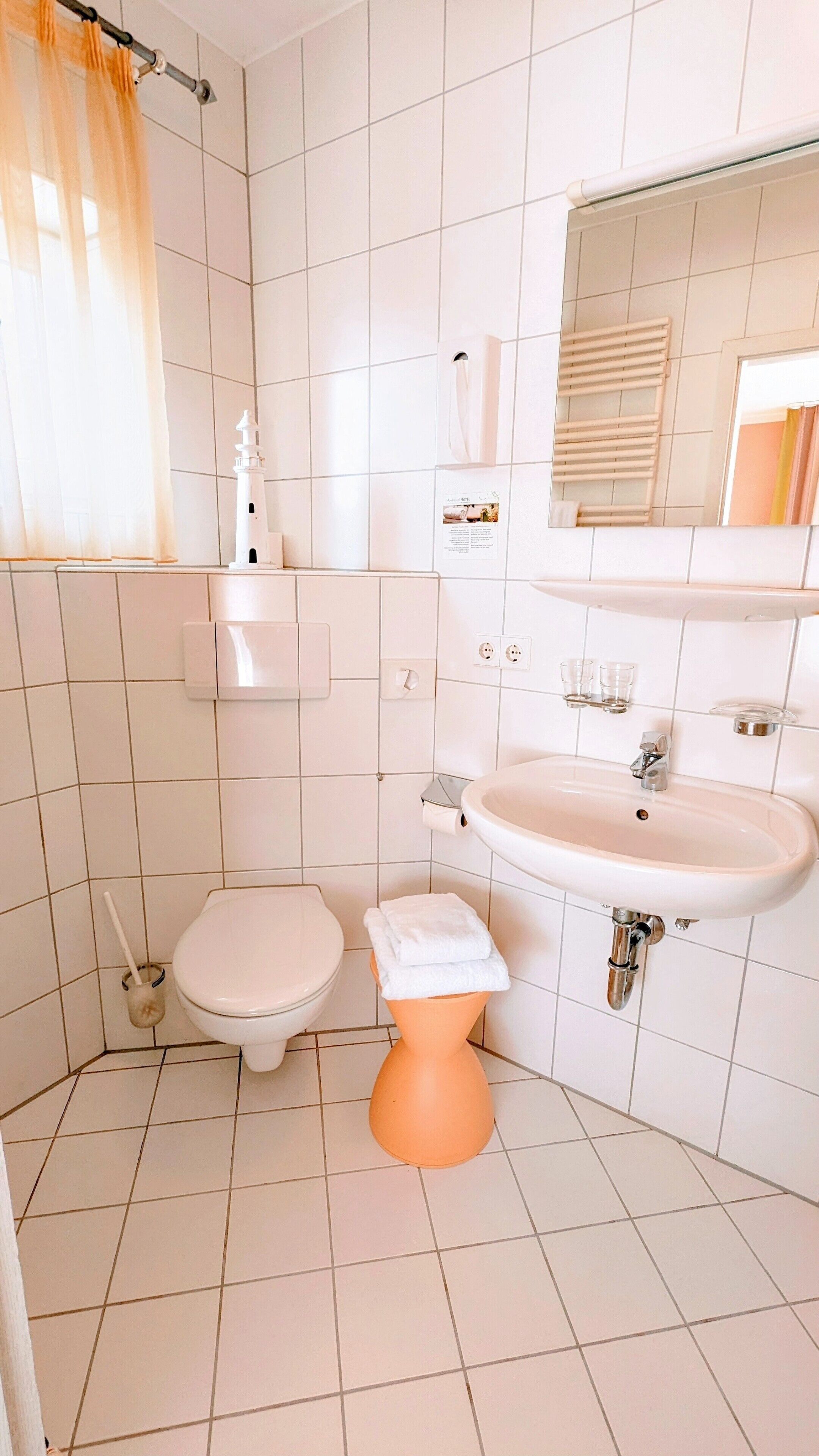 Ein Badezimmer mit weiß gefliesten Wänden, einer weißen Toilette, einem Standwaschbecken, einem Spiegel und einem Handtuchhalter.