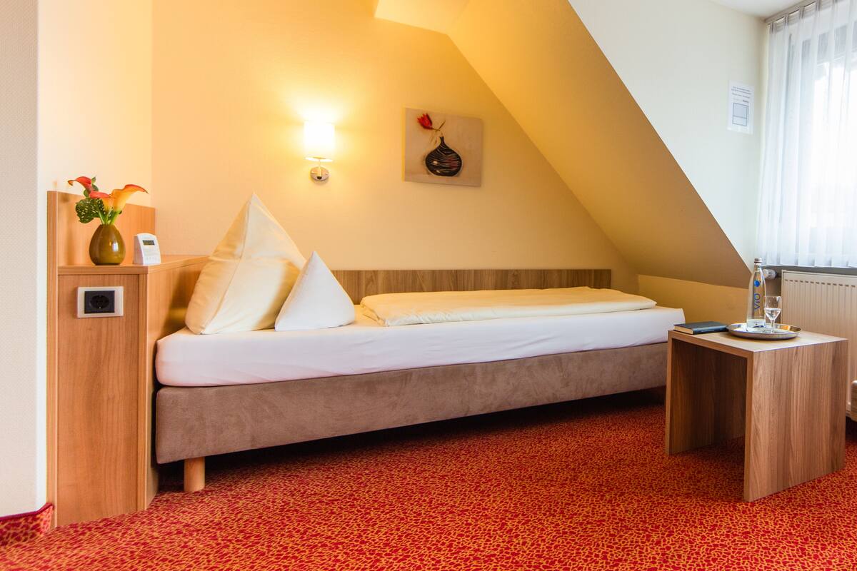 Une chambre d’hôtel avec un lit, une table de chevet, un vase de fleurs, une lampe fixée au mur et une fenêtre avec des rideaux.