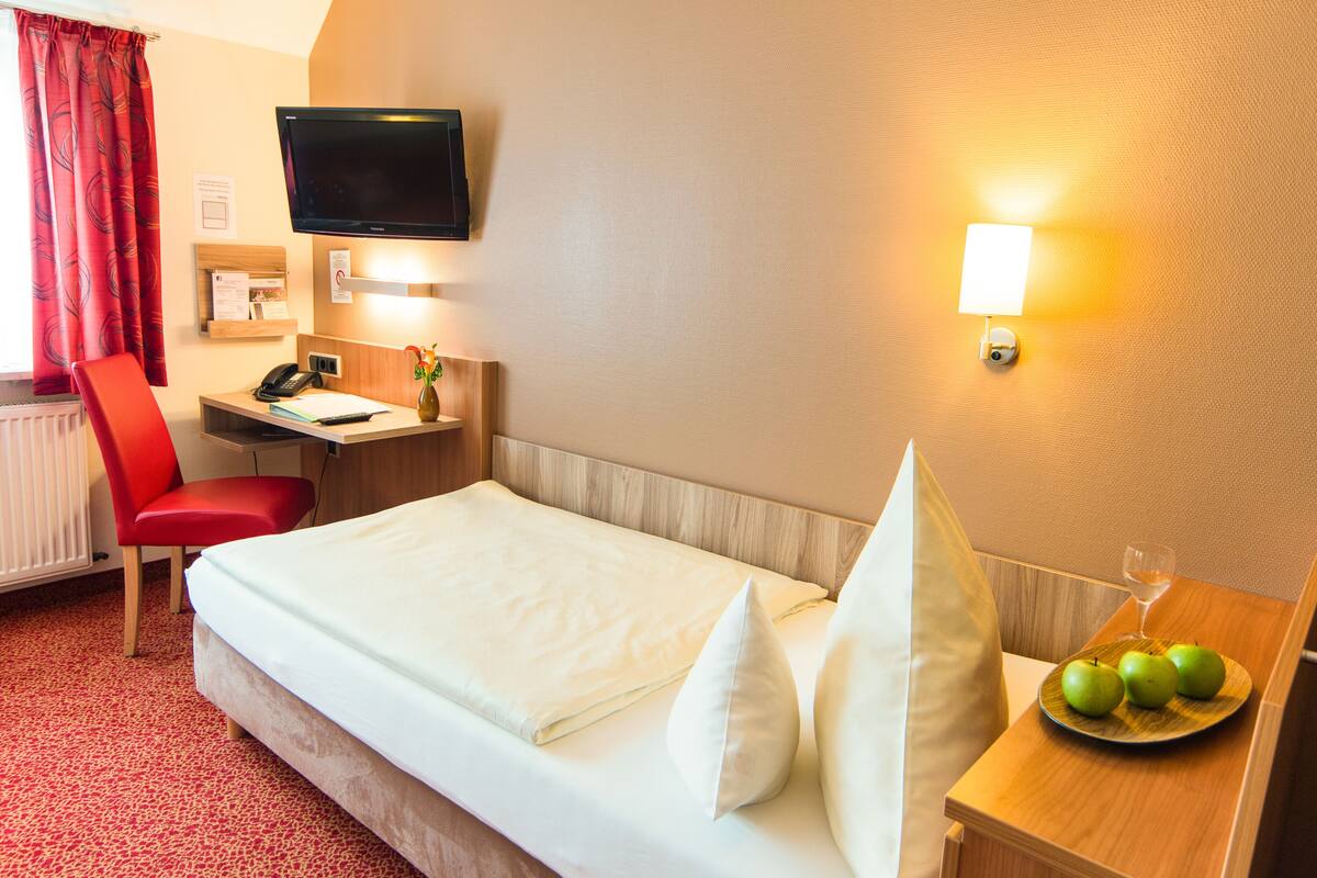 Une chambre d’hôtel avec un lit, un bureau, une chaise, une télévision et une table de chevet sur laquelle se trouvent des pommes et un verre d’eau.