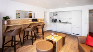 A modern living room with a wooden floor, a round ottoman, a coffee table, and a sofa with a red cushion. There is a kitchen area with white cabinets and a countertop. - Ambient Hotel Am Europakanal (Fuerth)