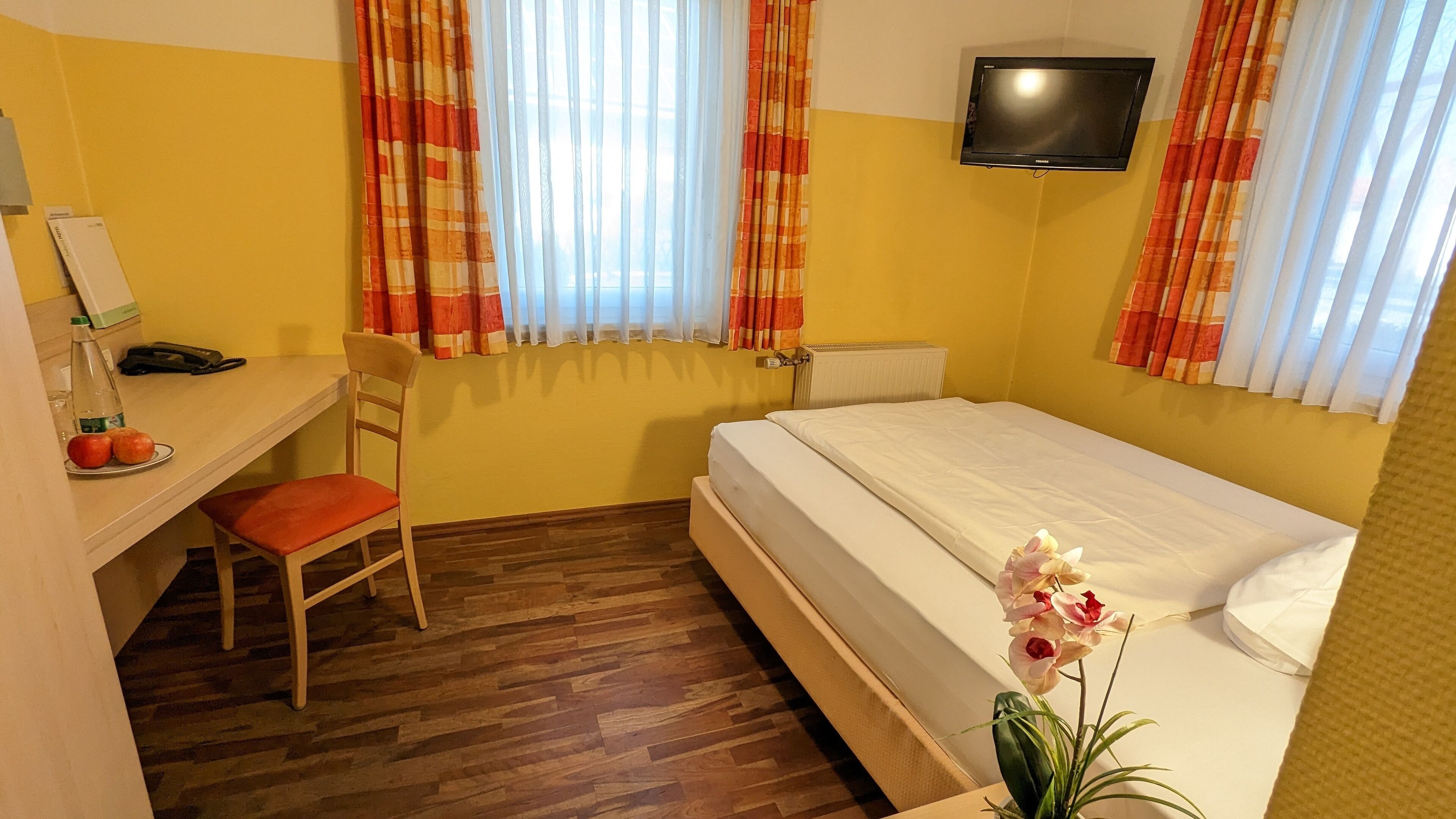 A hotel room with a wooden floor, a desk with a chair, a bed with white bedding, a television on the wall, and a window with orange curtains.