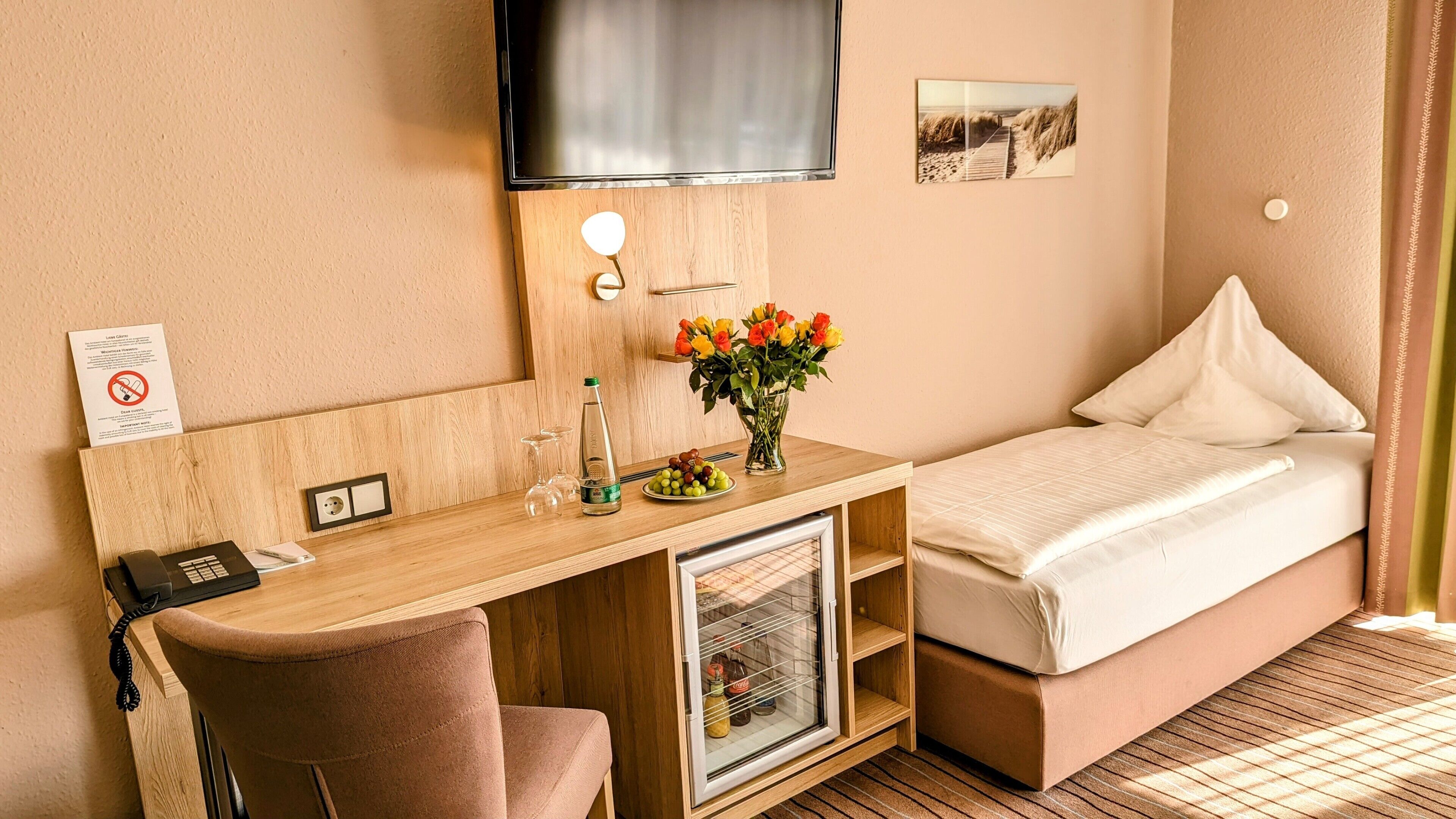 A compact hotel room with a bed, a desk with a chair, a TV mounted on the wall, and a small refrigerator.