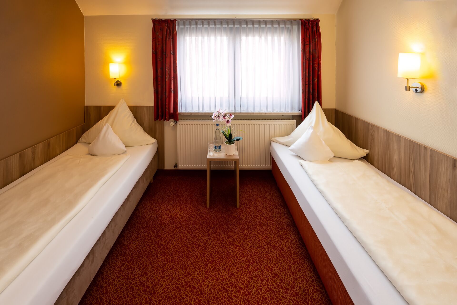 Une chambre d’hôtel avec deux lits simples, une petite table ornée d’un vase de fleurs et une fenêtre avec des rideaux.