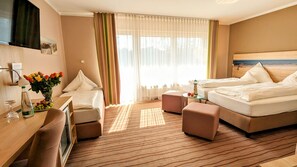 Ein Hotelzimmer mit zwei Betten, einem Schreibtisch, einem Stuhl, einem kleinen Tisch mit Blumenvase und einem großen Fenster mit Vorhängen.