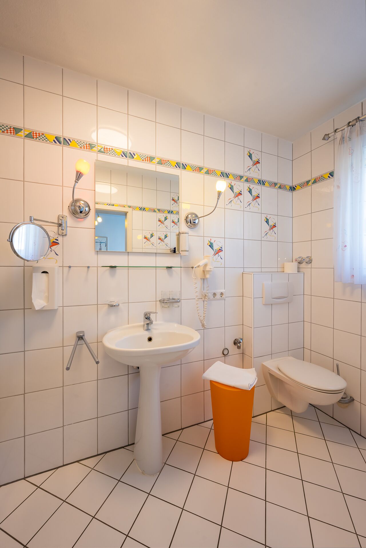 A bathroom with white tiled walls, a white sink, an orange bidet, and a toilet. There are two mirrors, one above the sink and one above the toilet.
