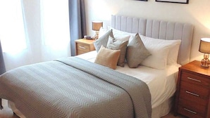 Un lit bien fait, recouvert d’une couvre-lit à motifs gris et blancs, avec une tête de lit rembourrée grise et deux tables de chevet dotées de lampes.