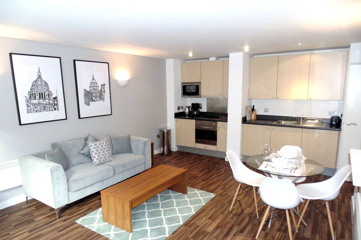 A modern living room with a sofa, coffee table, and dining area with chairs. There is a kitchen with cabinets and appliances. Two framed pictures are on the wall.
