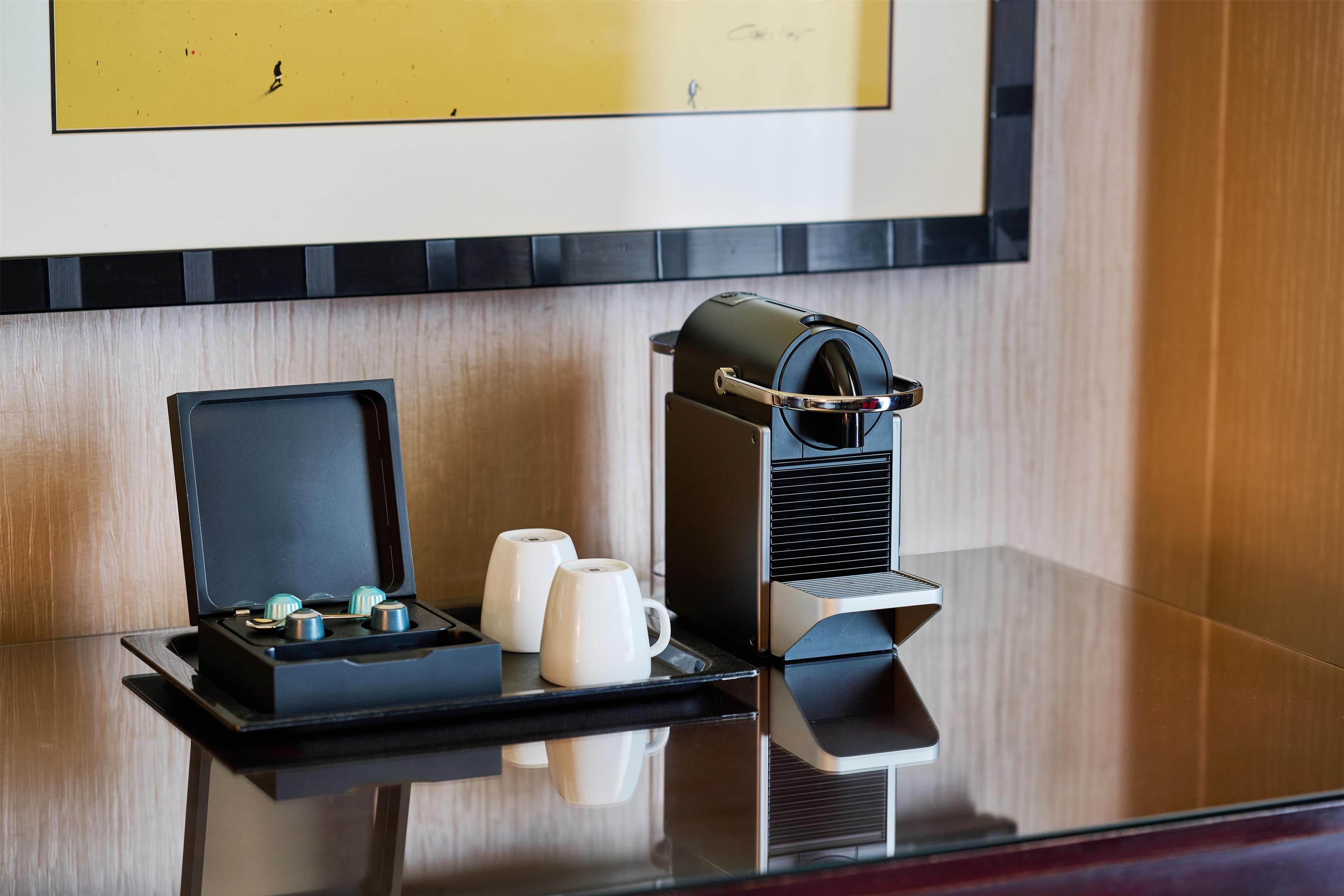 Máquina de café com bandeja, duas chávenas brancas e uma pequena caixa sobre uma superfície refletora.