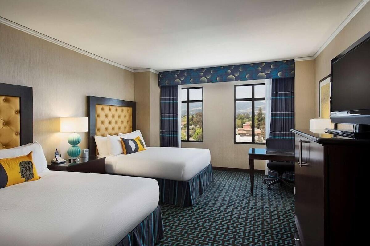 A hotel room with two beds, a desk, a TV, and large windows with a view.
