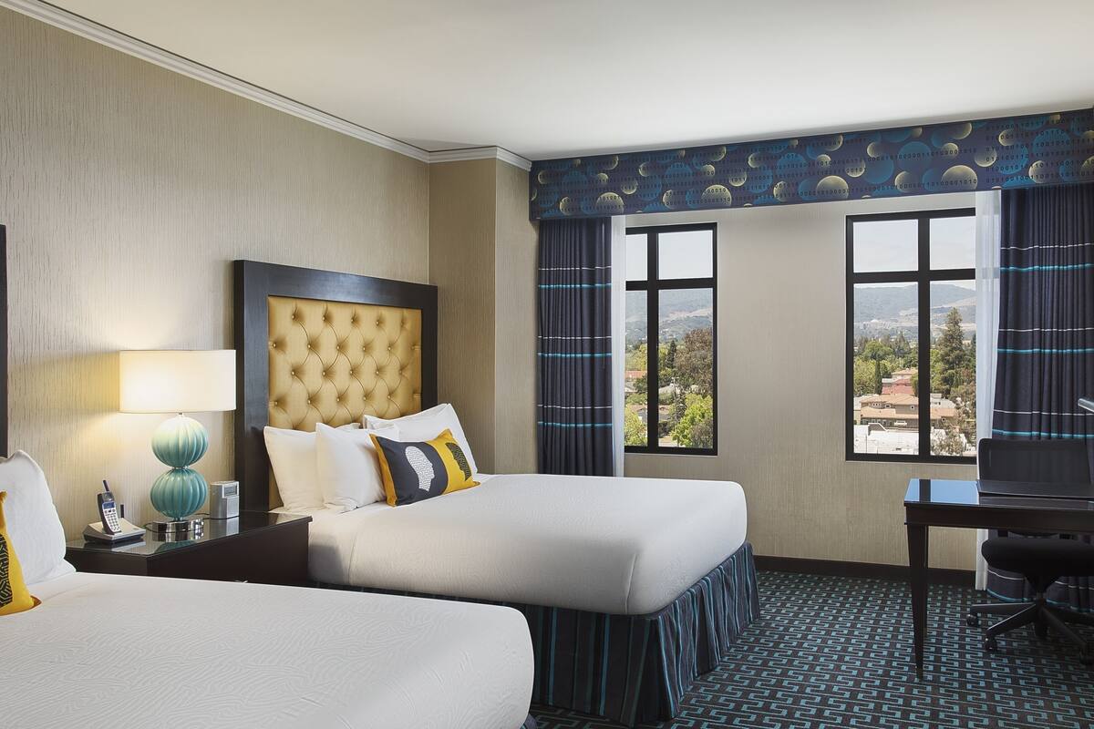 A hotel room with two beds, a desk, a chair, a lamp, and a view of the outside.
