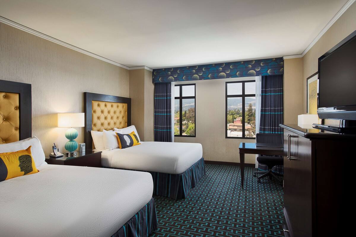 A hotel room with two beds, a desk, a TV, and a view of the outdoors.