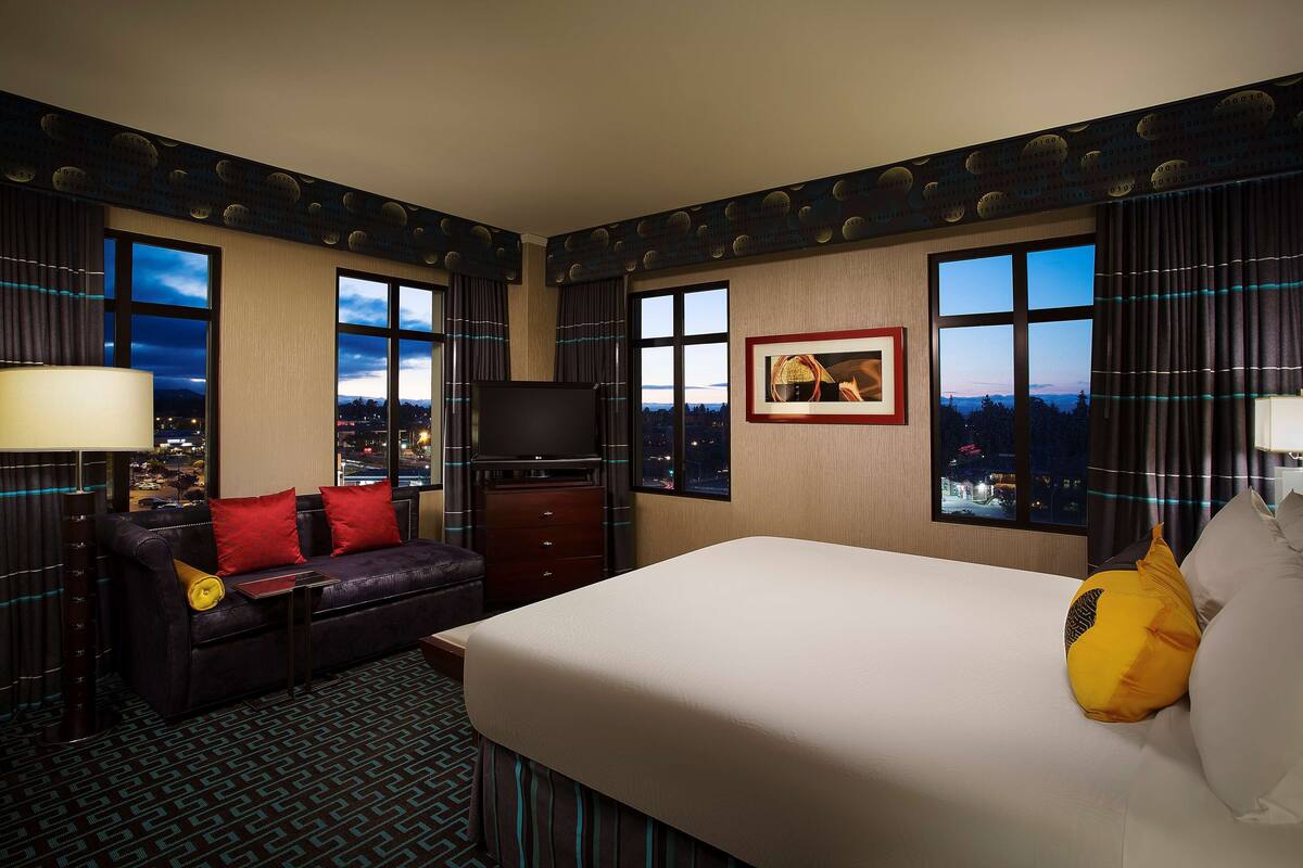 A hotel room with a large bed, a sofa with red cushions, a television, and a city view through the windows.