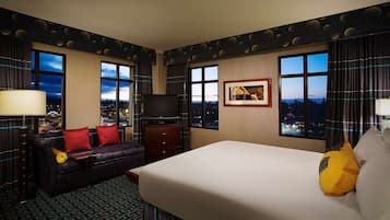 Ein Hotelzimmer mit einem großen Bett, einer Couch mit roten Kissen, einem Fernseher und Blick auf die Stadt durch die Fenster.