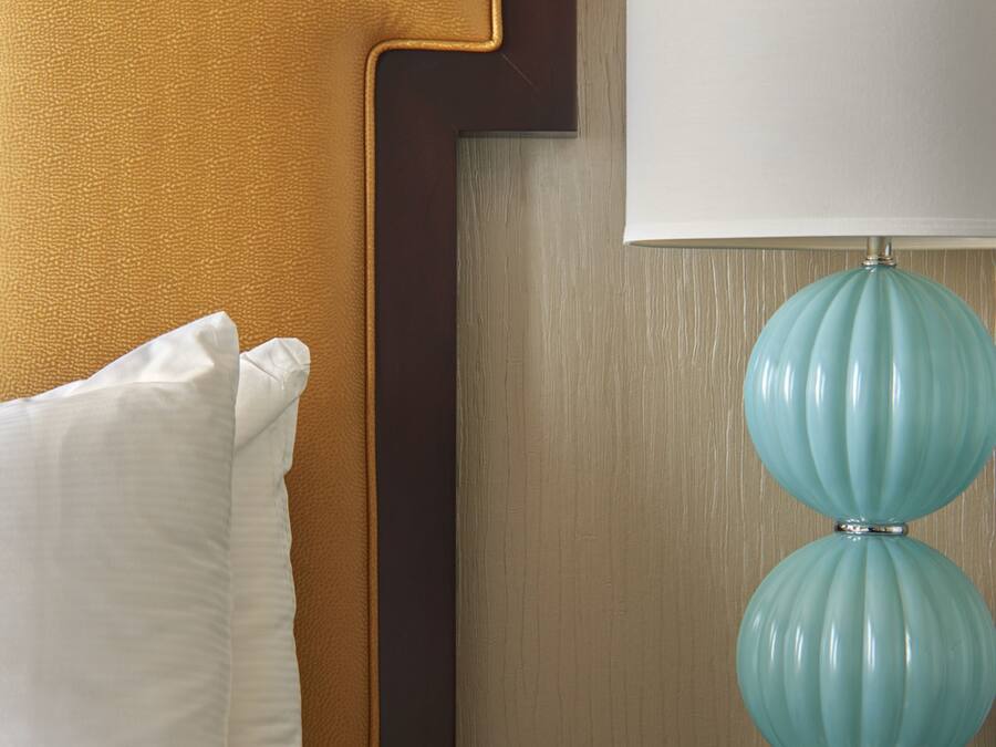 A neatly made bed with white linens and a single white pillow, a dark wooden nightstand with a teal lamp, and a wall with a textured wallpaper.