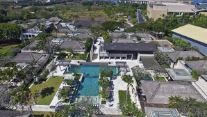 Vista aérea de um resort com uma piscina central, rodeada por vilas com telhados de palha, e vista para o oceano.