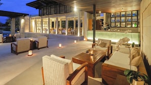 Un área de estar al aire libre con mobiliario de mimbre y velas, un bar con una vitrina de vidrio y una piscina al fondo.