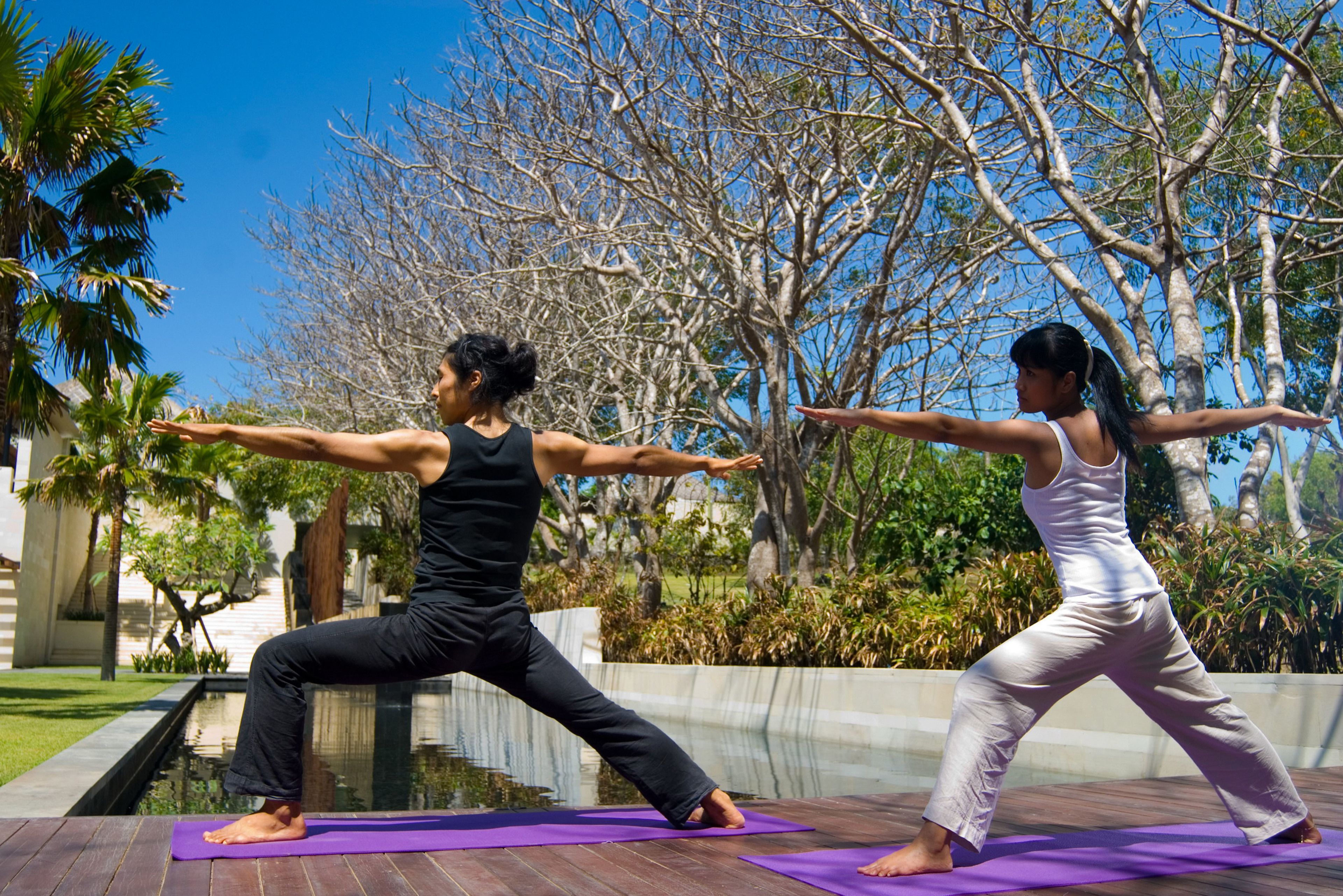 Dos personas practicando yoga al aire libre sobre una terraza de madera, con una piscina y árboles al fondo.