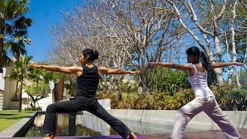 Dos personas practicando yoga al aire libre sobre una terraza de madera, con una piscina y árboles al fondo.