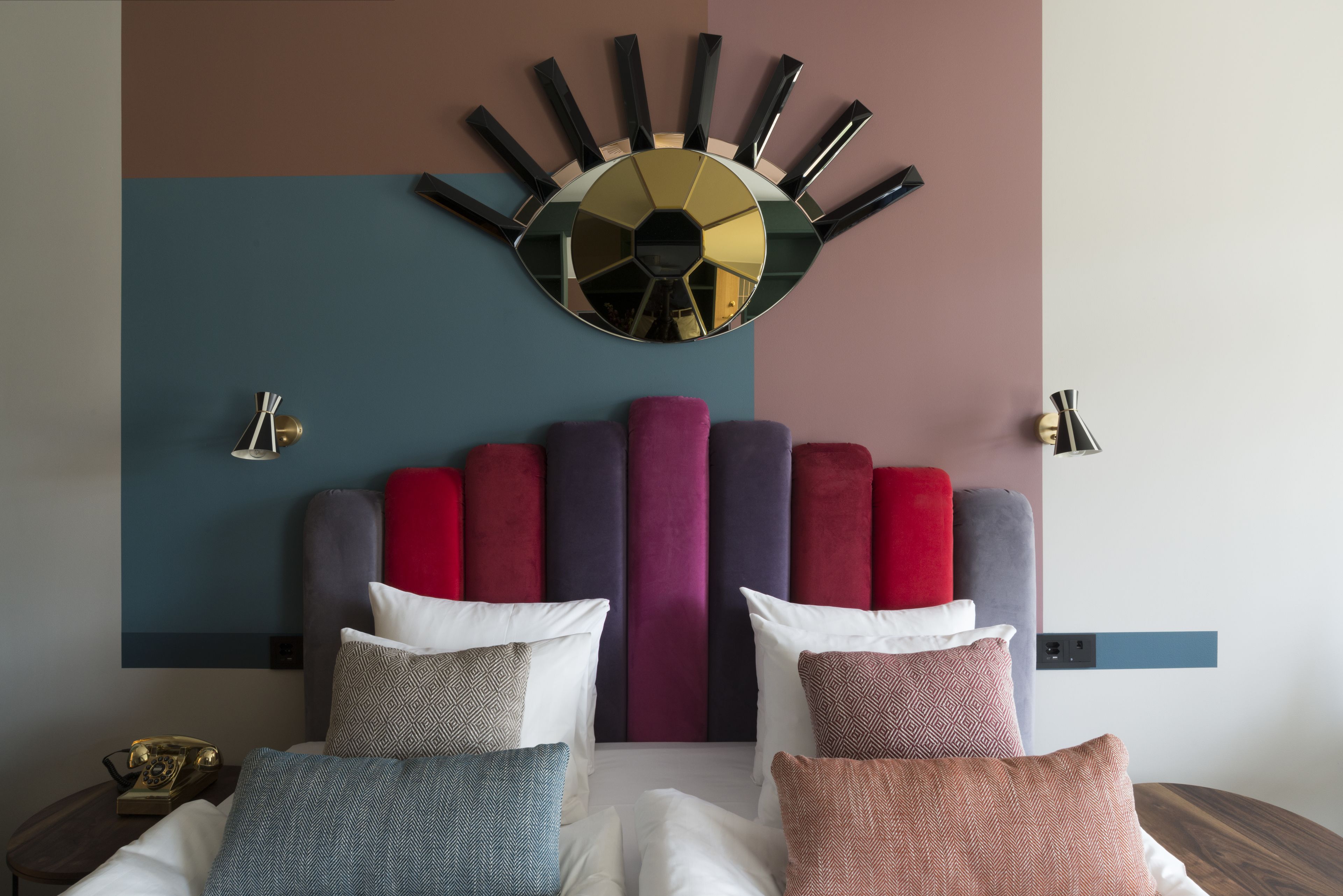 A modern hotel room with a large round mirror, a tufted headboard, and a variety of patterned pillows.