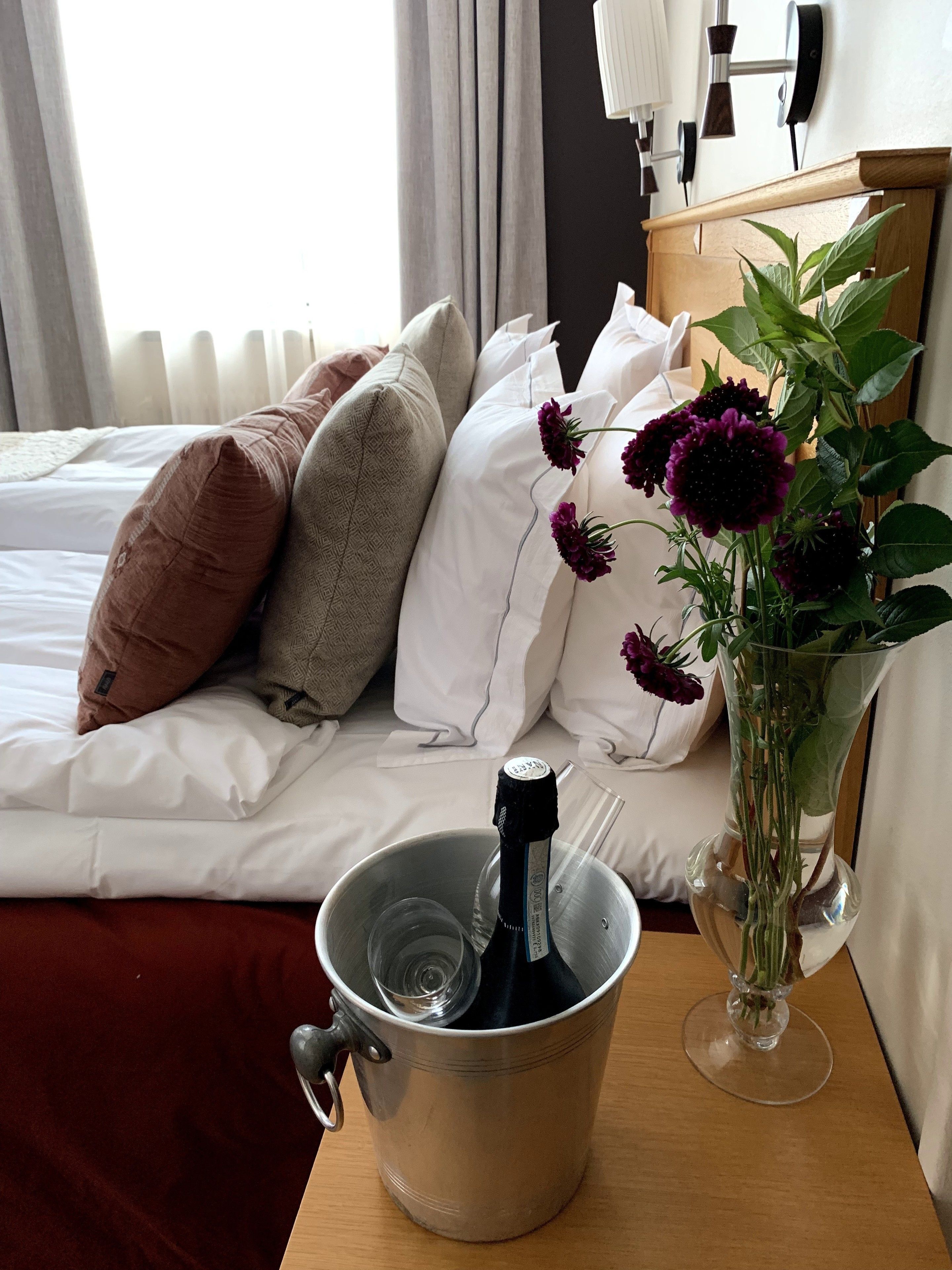 Habitación de hotel con cama, almohadas, un florero con flores y un cubo con una botella y vasos.