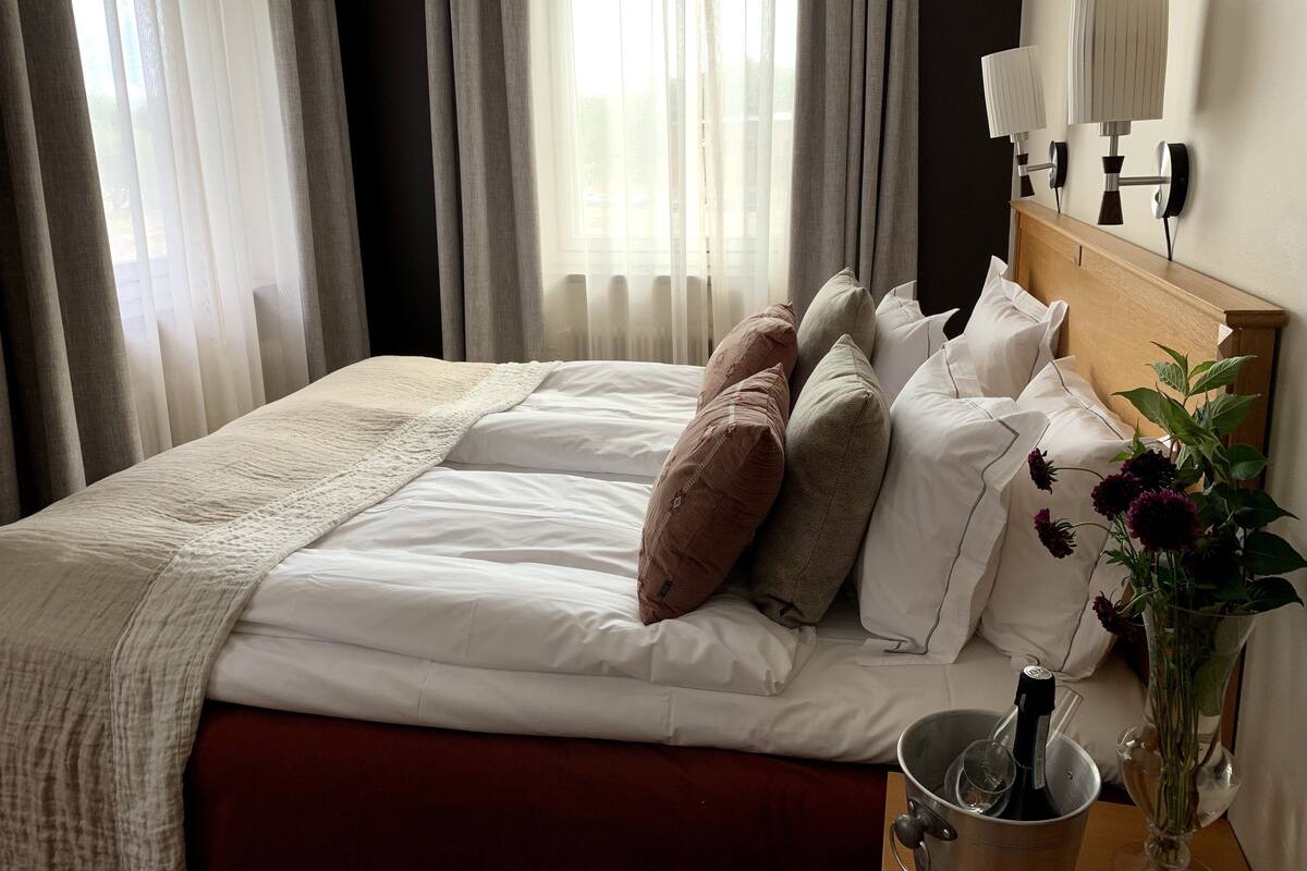 A neatly made bed with white linens and multiple pillows, a bedside table with a bottle and a vase of flowers, and a window with sheer curtains allowing natural light.