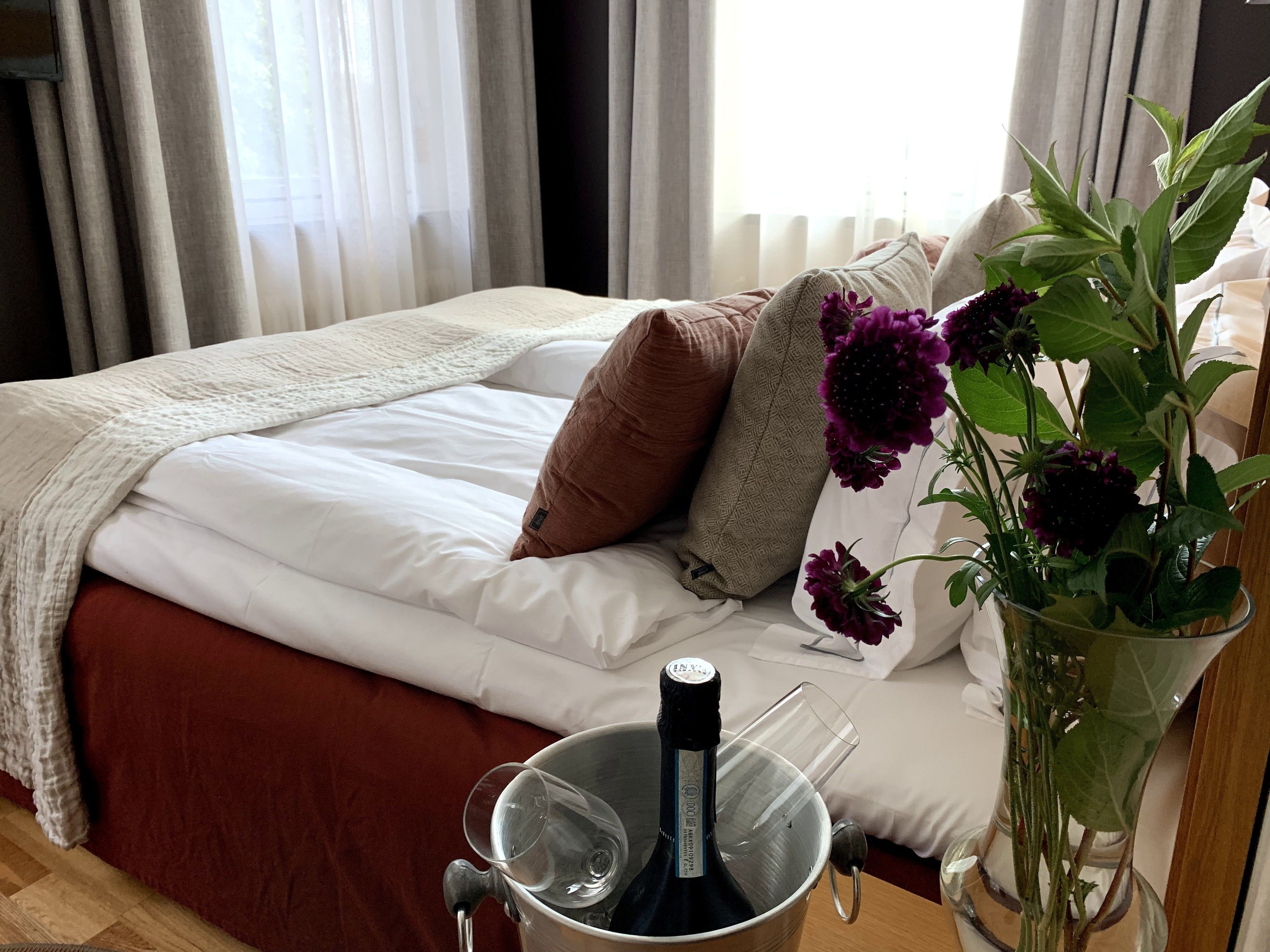 Una cama bien hecha con edredón blanco y almohadas marrones, una copa de vino en un cubo, y un florero con flores moradas sobre una mesa de madera.