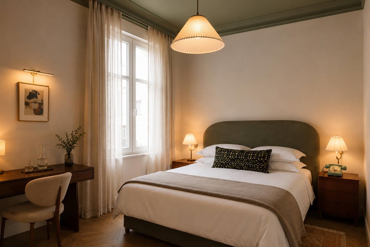 A hotel room with a bed, bedside lamps, a desk with a chair, a window with curtains, and a wall-mounted picture.