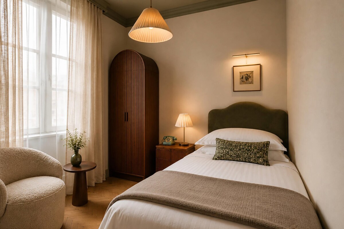 A hotel room with a bed, a chair, a bedside table with a lamp, a wardrobe, and a window with curtains.