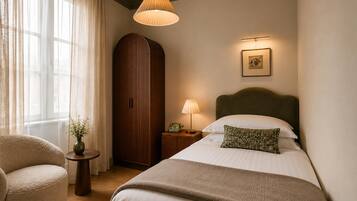 Une chambre d’hôtel comprenant un lit, une chaise, une table de chevet avec une lampe, une armoire et une fenêtre avec des rideaux.