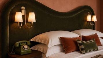 A hotel room with a classic design, featuring a curved headboard, a vintage rotary phone, and a neatly made bed with multiple pillows.