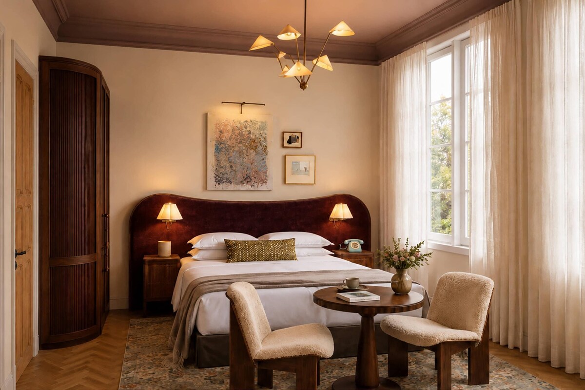 A hotel room with a large bed, two bedside lamps, a chandelier, a round table with a book and a cup, a chair, and a window with curtains.
