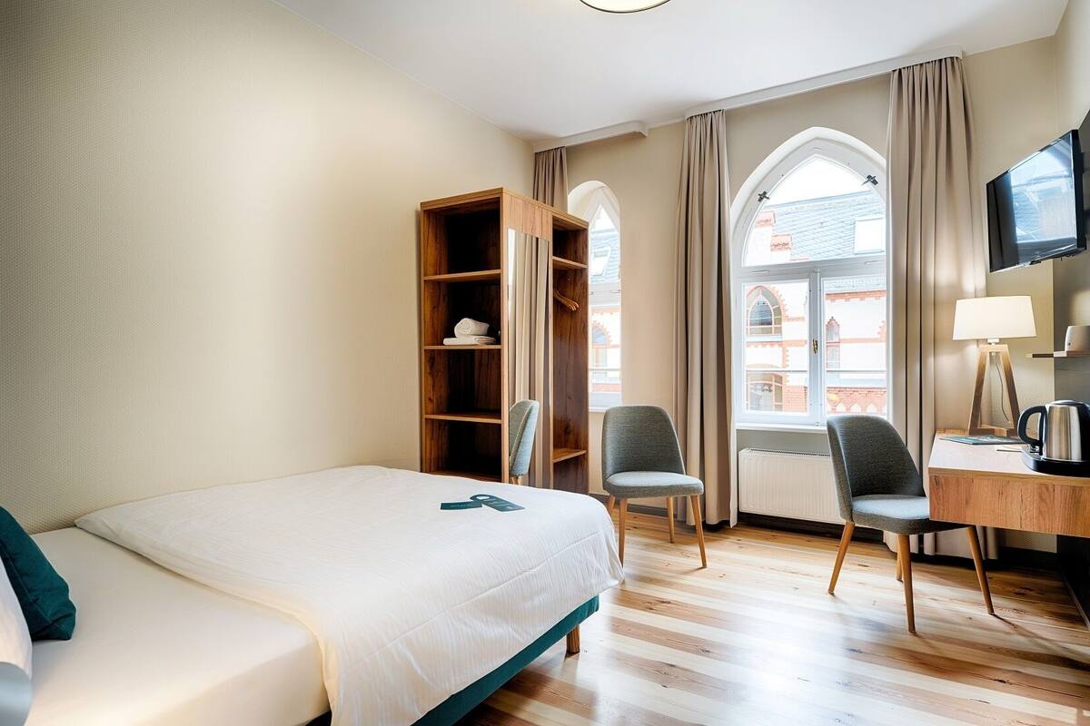Une chambre d’hôtel comprenant un lit, deux chaises, un bureau avec une lampe, une télévision et une fenêtre avec des rideaux.