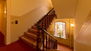 Un escalier en bois avec une rampe, un sol recouvert d’une moquette rouge, une fenêtre avec une plante et un luminaire fixé au mur.