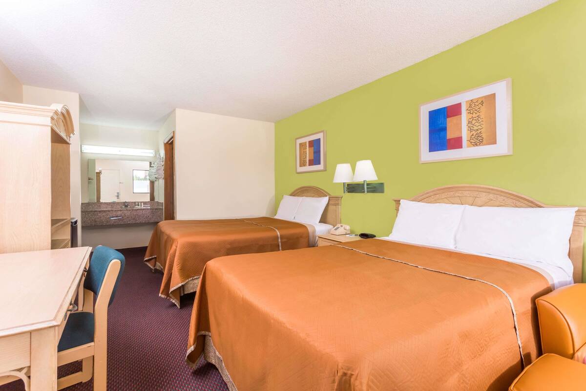 A hotel room with two beds, a desk, and a chair. There is a bathroom visible in the background.