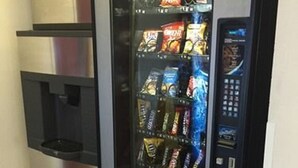 A vending machine with snacks and beverages, located in a corner with a tiled floor. - Osceola Grand Hotel (Evart)