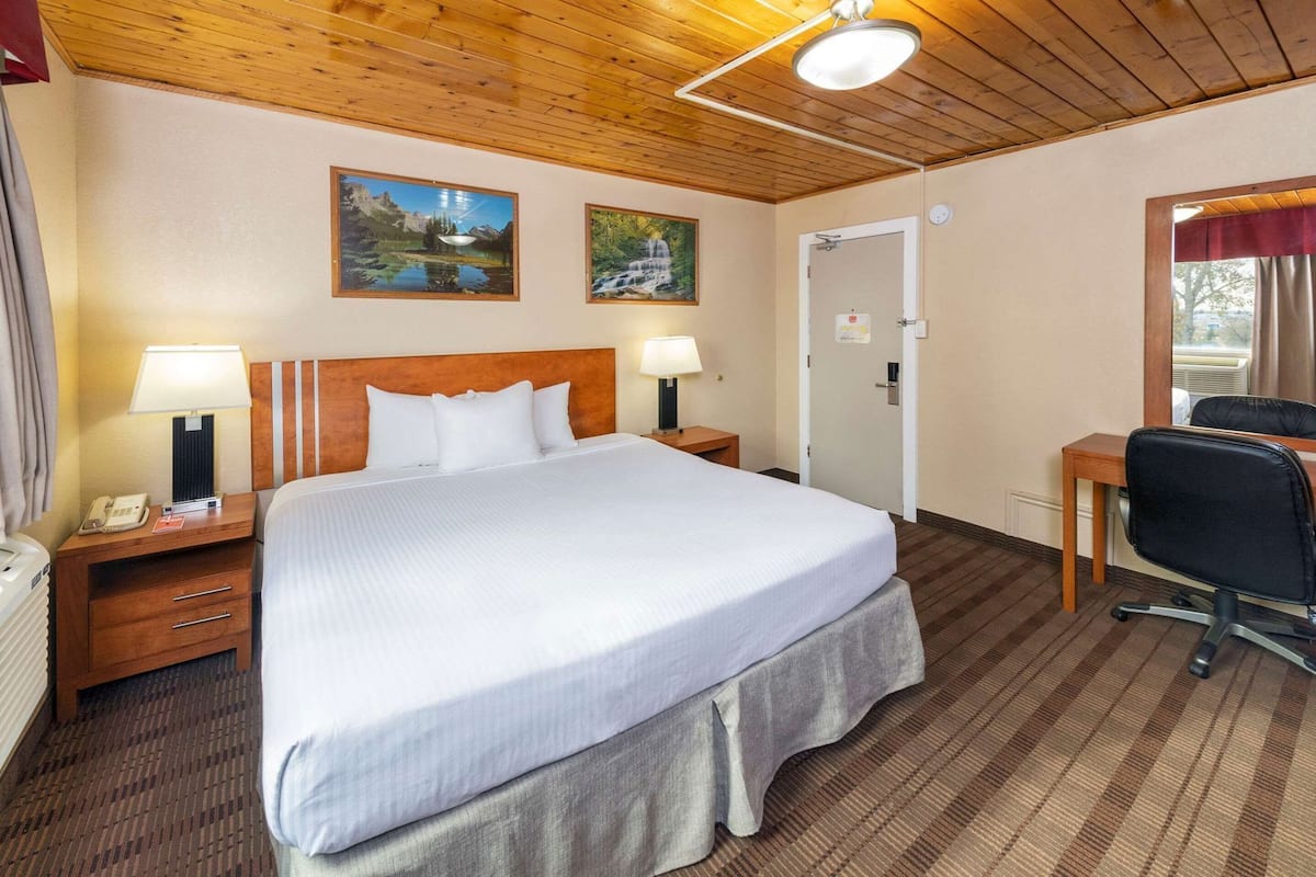 A hotel room with a large bed, two bedside tables with lamps, a desk with a chair, and two framed pictures on the wall.