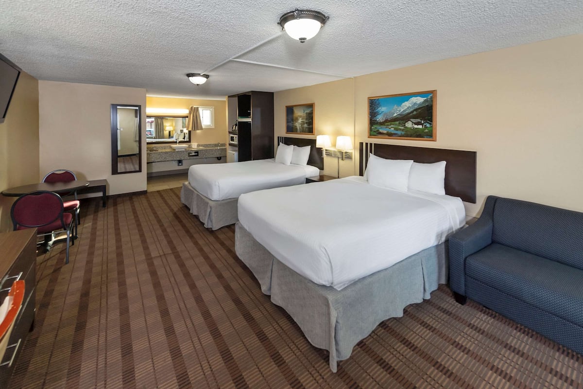 A hotel room with two beds, a desk, a chair, a sofa, a bathroom, and a painting on the wall.