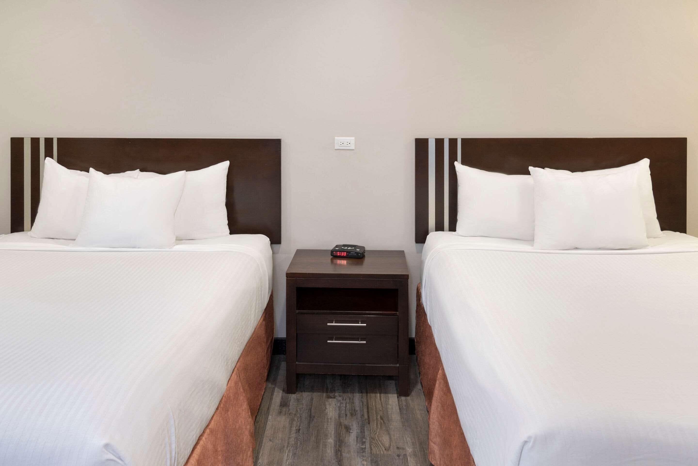 Habitación de hotel con dos camas, cada una con ropa de cama blanca y cabeceros oscuros. Entre las camas hay una mesita de noche de madera con un reloj encima.