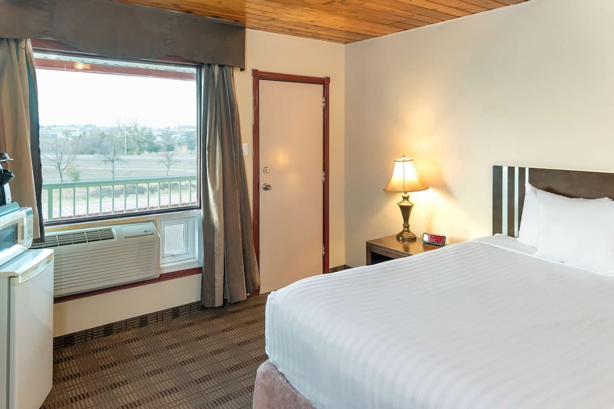 A hotel room with a bed, a window with curtains, a lamp, a clock, and a radiator.