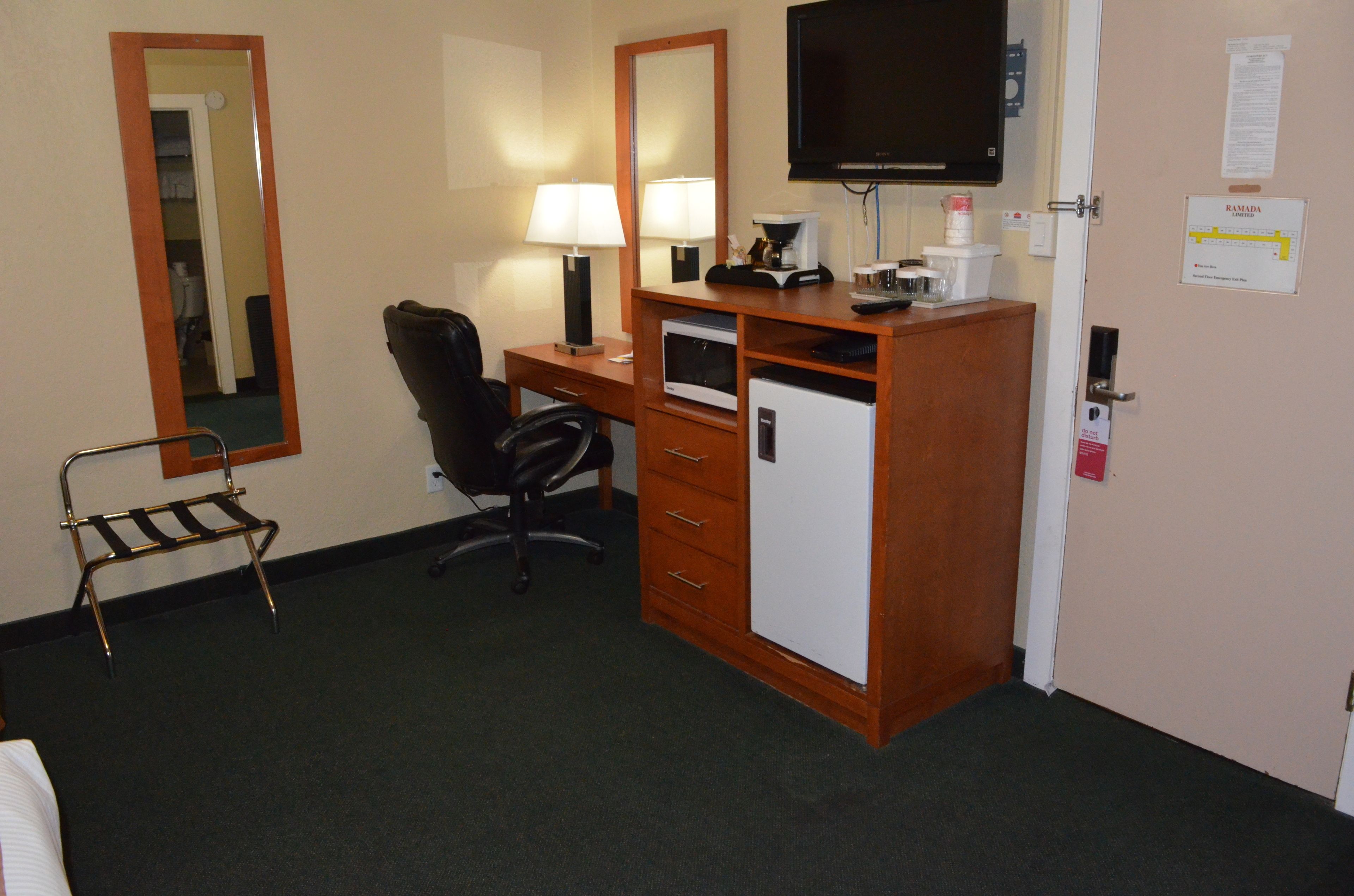 A hotel room with a desk, chair, small refrigerator, and a television mounted on the wall.
