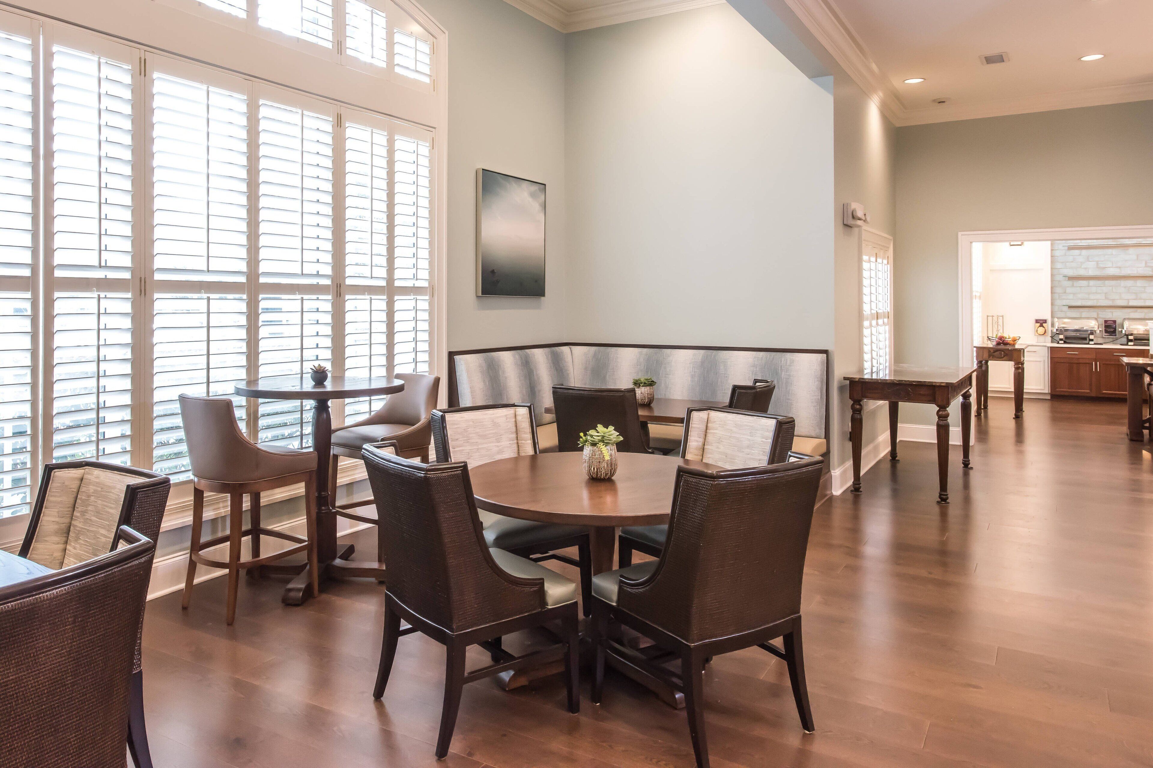 A dining area with a round table and chairs, large windows with blinds, and a kitchen in the background.