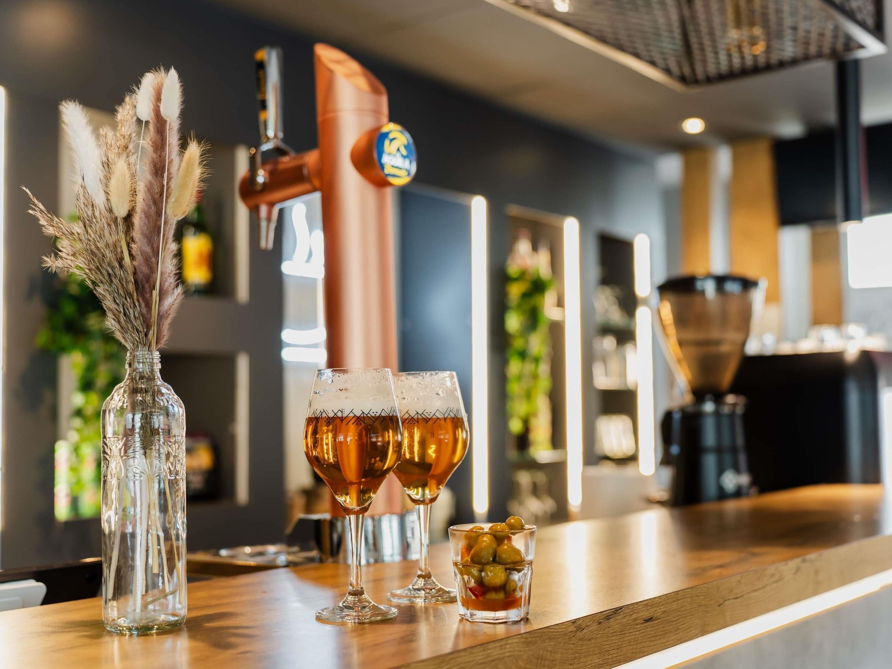 Una zona de bar con un grifo de cobre, dos vasos de cerveza, un vaso con aceitunas y un jarrón decorativo con plumas de miscania.