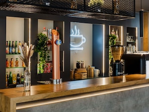 A bar area with a copper tap, shelves of bottled beverages, a neon coffee cup sign, and a coffee machine. - ibis Barcelona Castelldefels (Castelldefels)