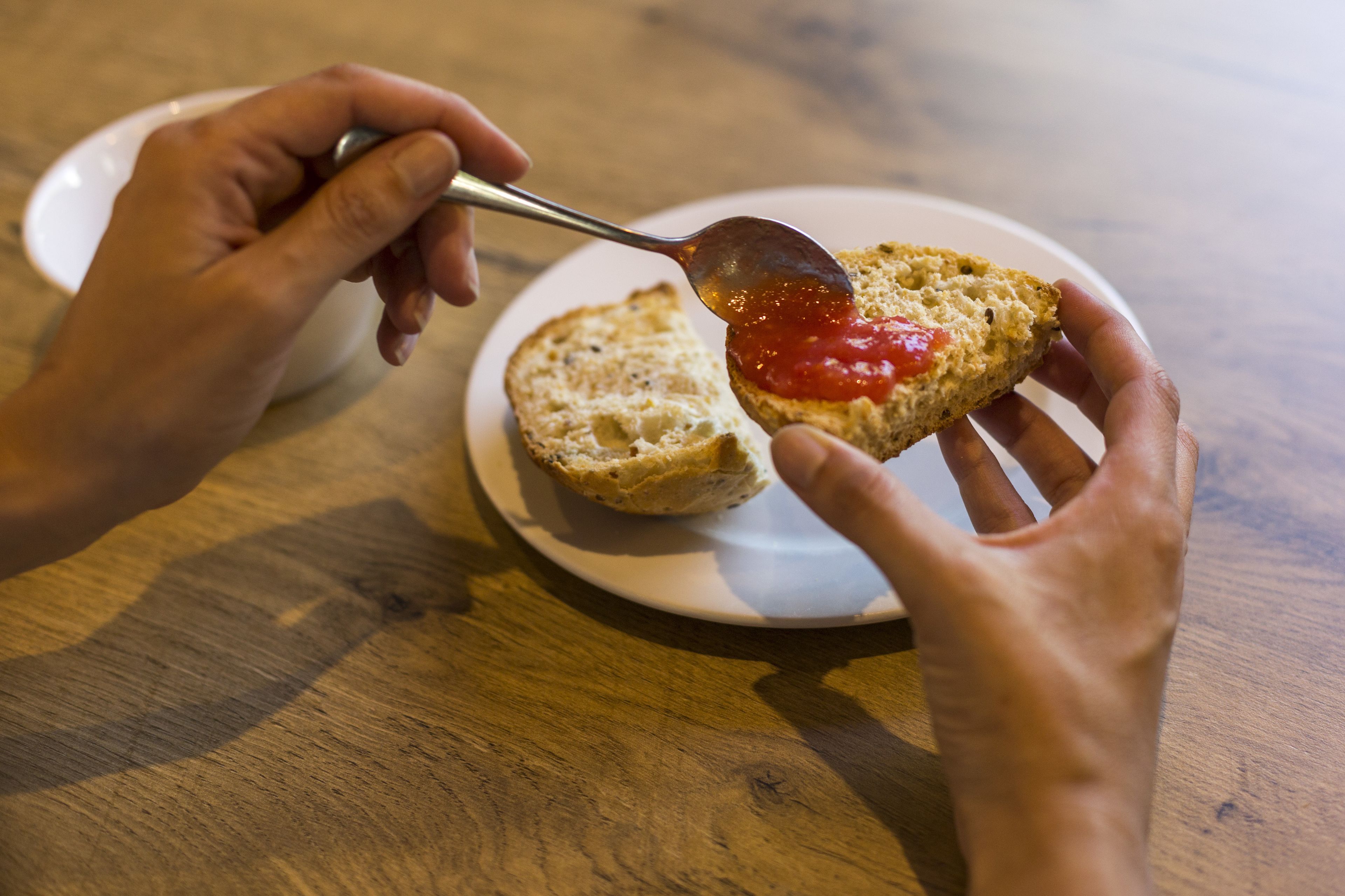 A person spreading jam on a slice of bread on a white plate.