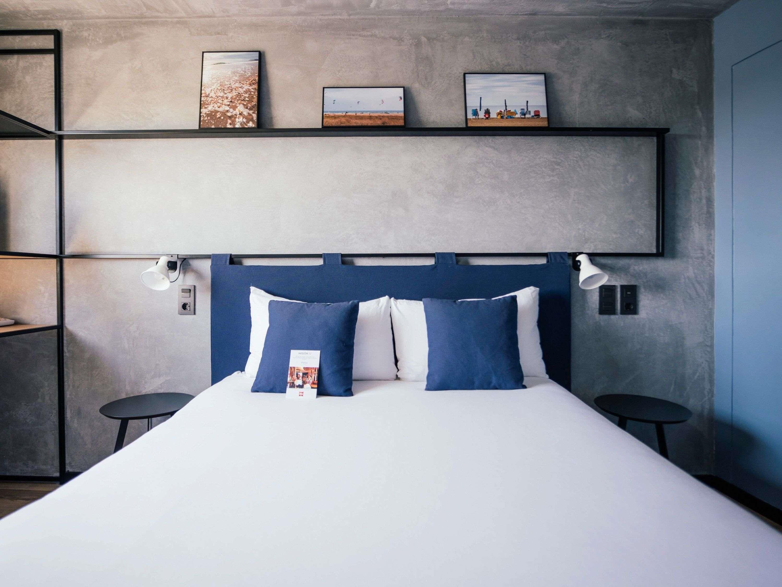 Quarto de hotel moderno com uma cama grande, cabeceira com iluminação embutida, uma prateleira com quadros e duas mesas de cabeceira.