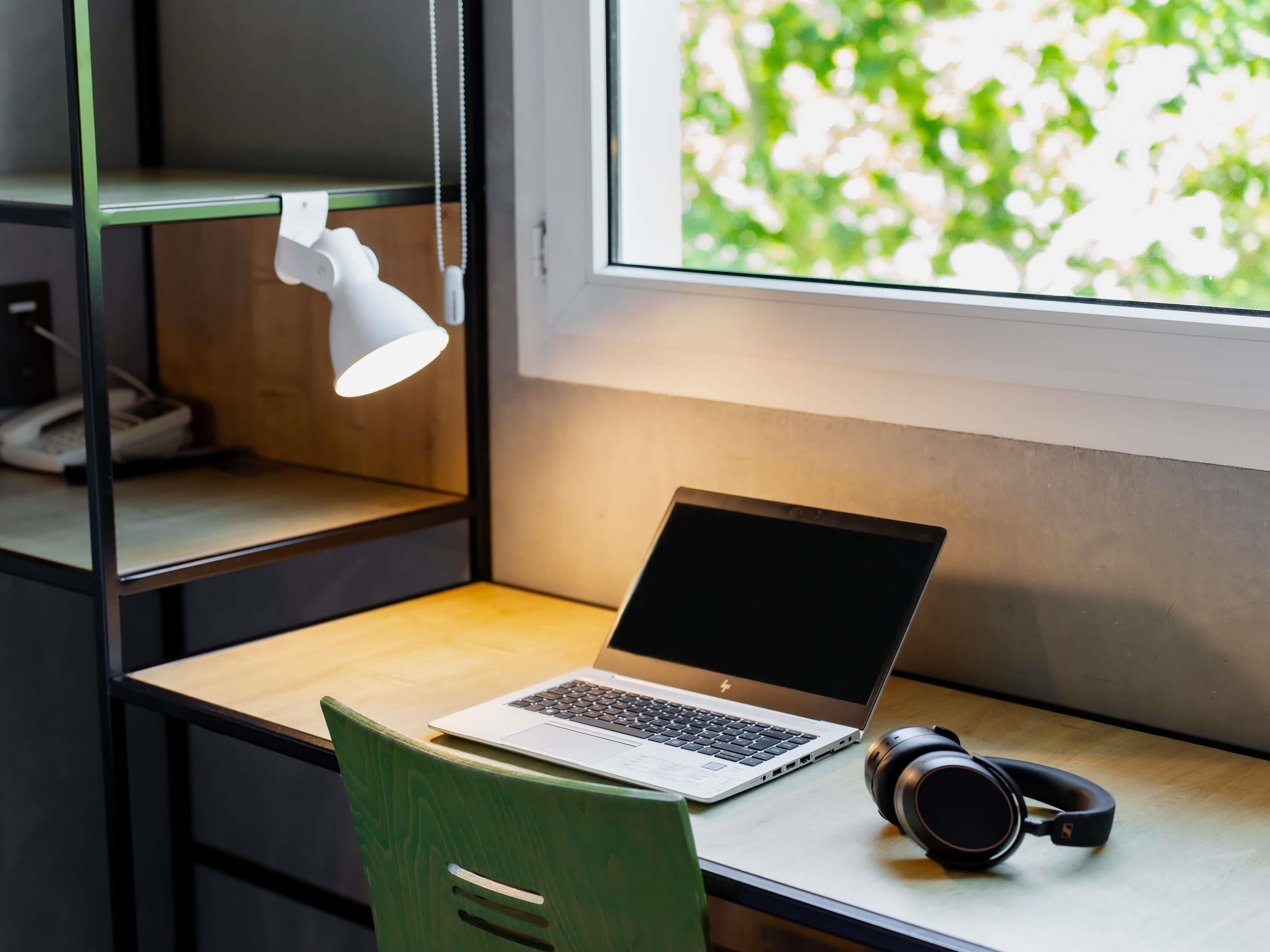Una laptop sobre un escritorio con auriculares, una lámpara de escritorio blanca y una ventana con vista a follaje.